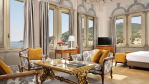 Habitación de hotel lujosa con una cama grande, un sofá, una mesa de comedor y un televisor. La habitación tiene ventanas amplias con vistas al mar y a las colinas.