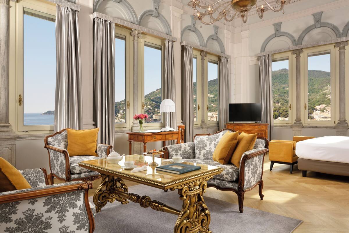 A luxurious hotel room with a large bed, a sofa, a dining table, and a TV. The room has large windows with a view of the sea and hills.