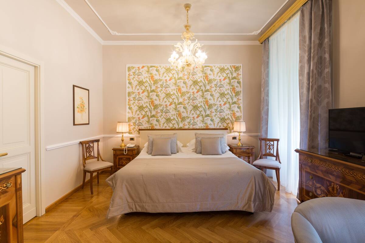 A hotel room with a large bed, two bedside tables with lamps, a television, and a patterned wallpaper.