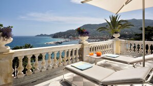 Un balcón con vista al mar, sillones de descanso, una mesa con bebidas y plantas decorativas.