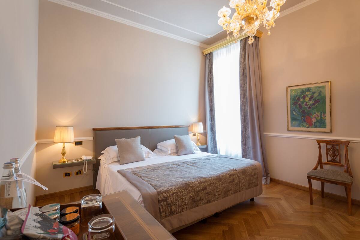 A neatly arranged hotel room with a large bed, bedside lamps, a wooden chair, and a painting on the wall.