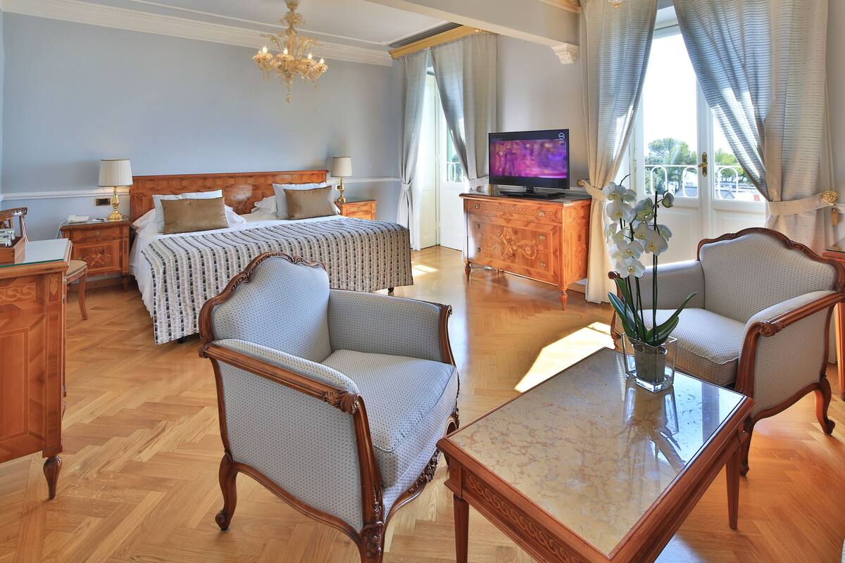 A hotel room with a double bed, two armchairs, a wooden dresser, a TV, and a large window with curtains.