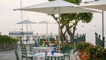 An outdoor dining area with tables and chairs, umbrellas, and a view of the sea.