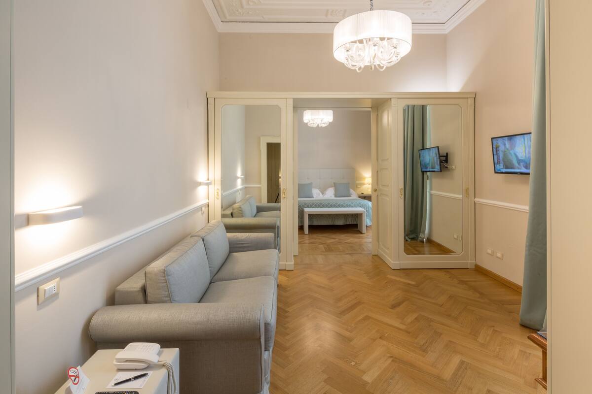 A hotel room with a sofa, a bedside table, a mirror, a chandelier, and a television.