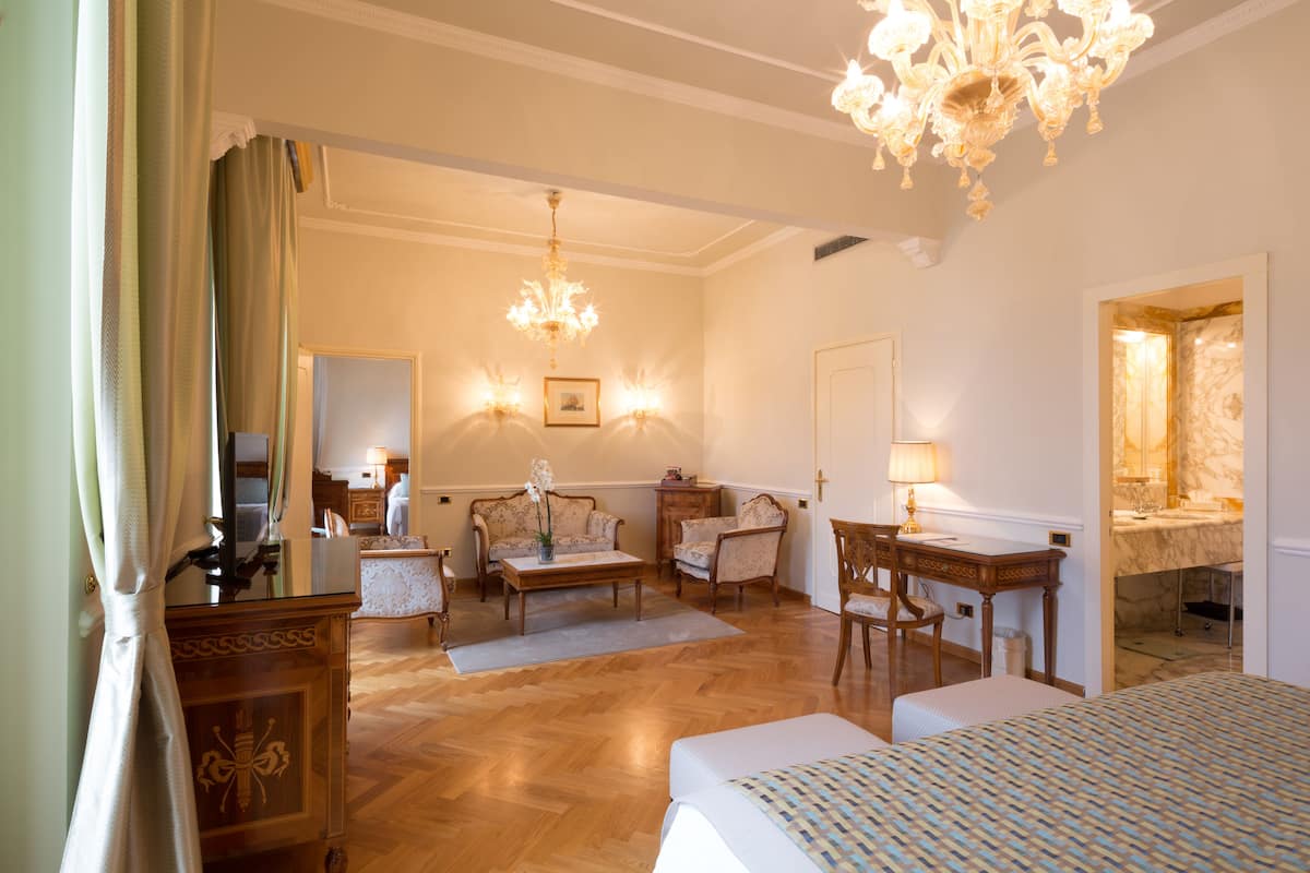 A hotel room with a large bed, a desk, two armchairs, a chandelier, and a bathroom visible through an open door.