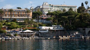Un hotel frente al agua con varias construcciones, un muelle con sillones de descanso y un litoral rocoso.