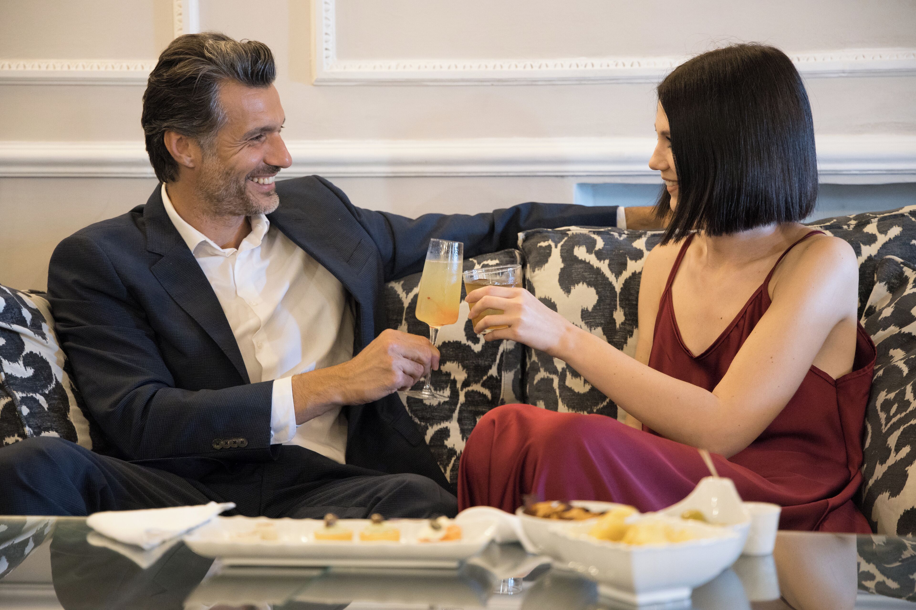 A man and a woman sitting on a patterned sofa, holding glasses of a beverage.