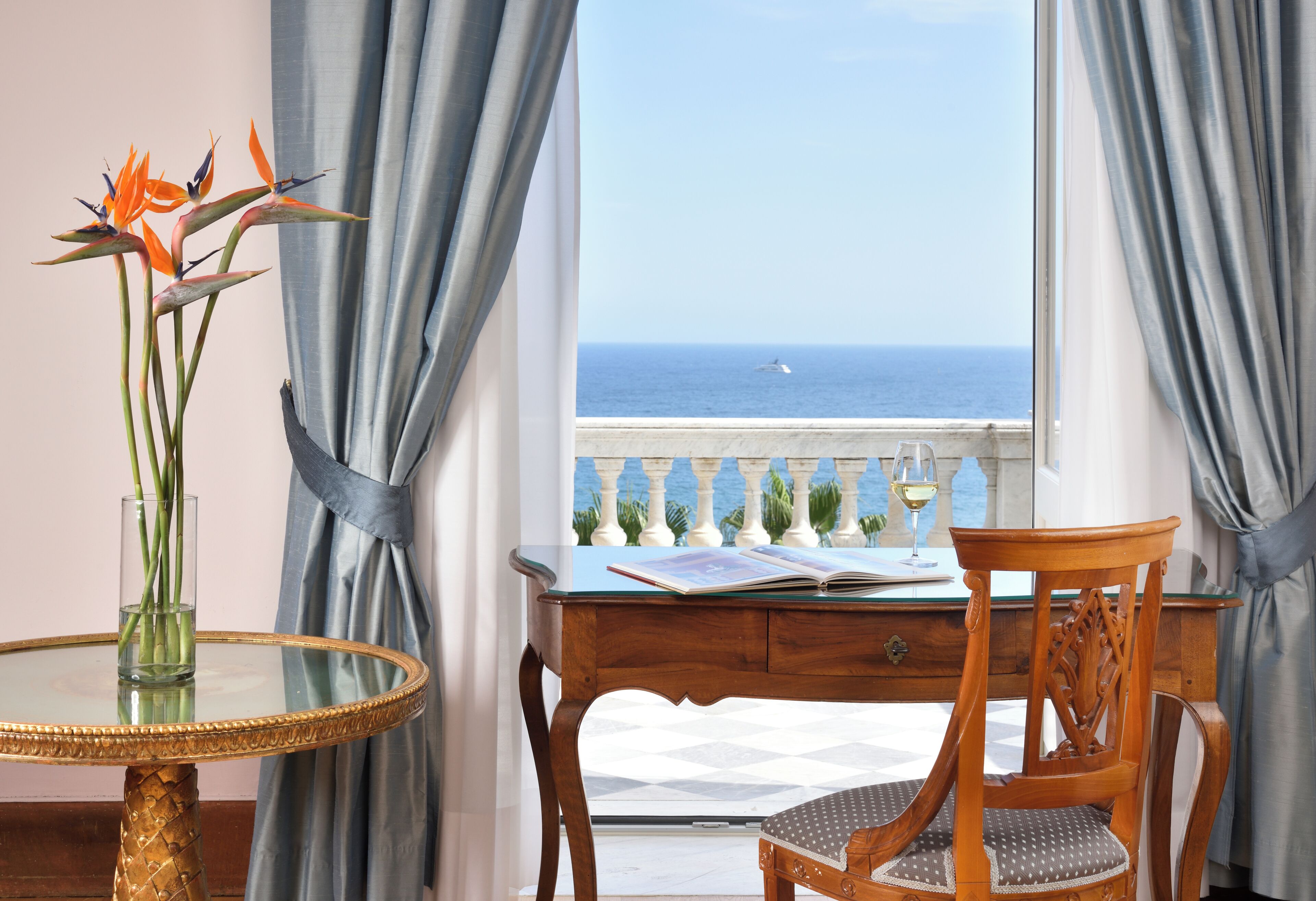 A room with a view of the sea, a table with a vase of flowers, and a chair with a book.