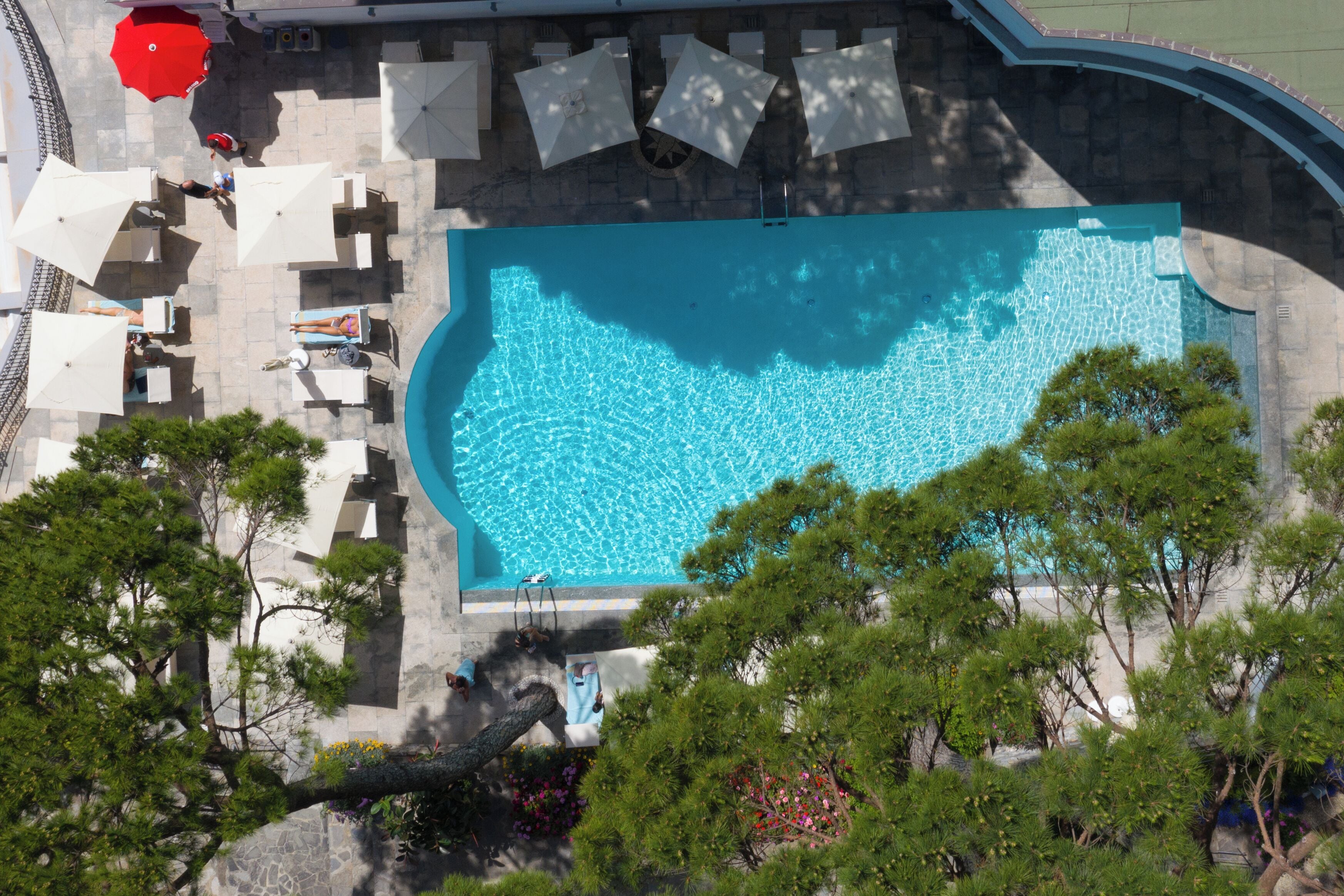 An aerial view of a swimming pool with surrounding lounge chairs and umbrellas, surrounded by trees and a stone wall.