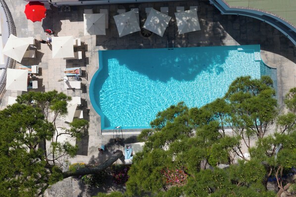 An aerial view of a swimming pool with surrounding lounge chairs and umbrellas, surrounded by trees and a stone wall.