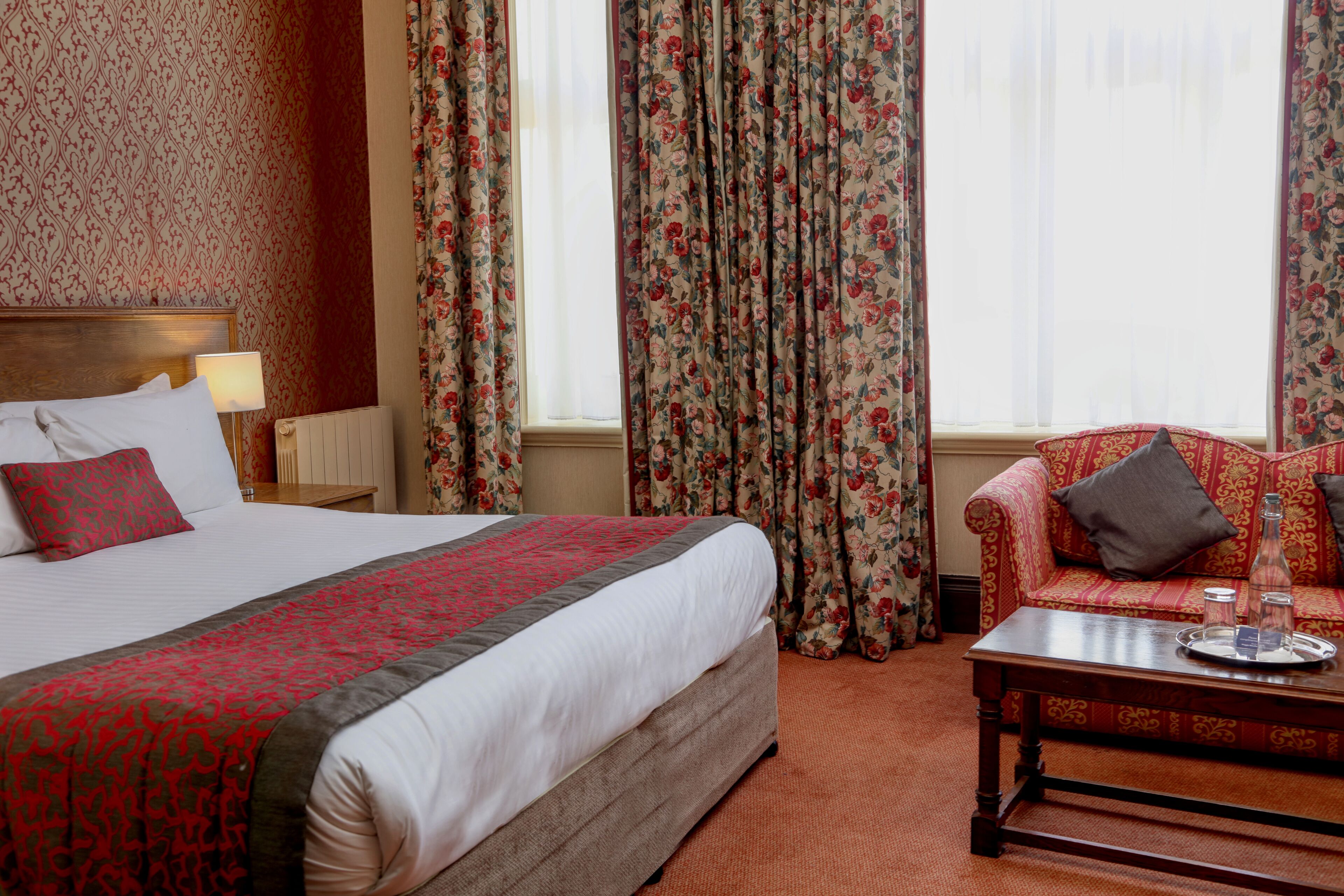 A hotel room with a large bed, a wooden headboard, a red and white patterned bedspread, a floral curtain, a red sofa with a grey pillow, and a wooden coffee table with a tray.
