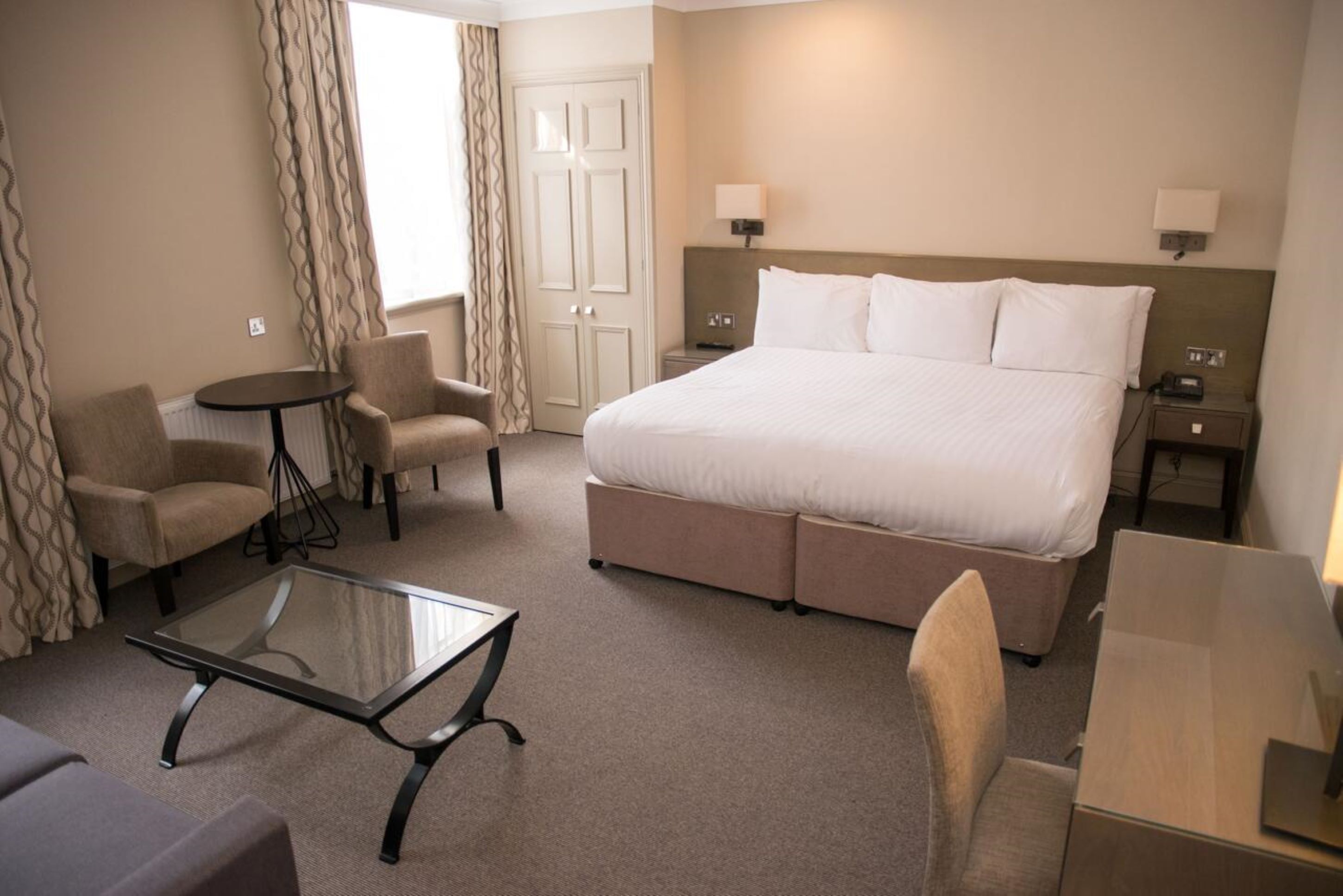 A hotel room with a large bed, two bedside tables, a glass coffee table, two armchairs, a sofa, and a door with curtains.