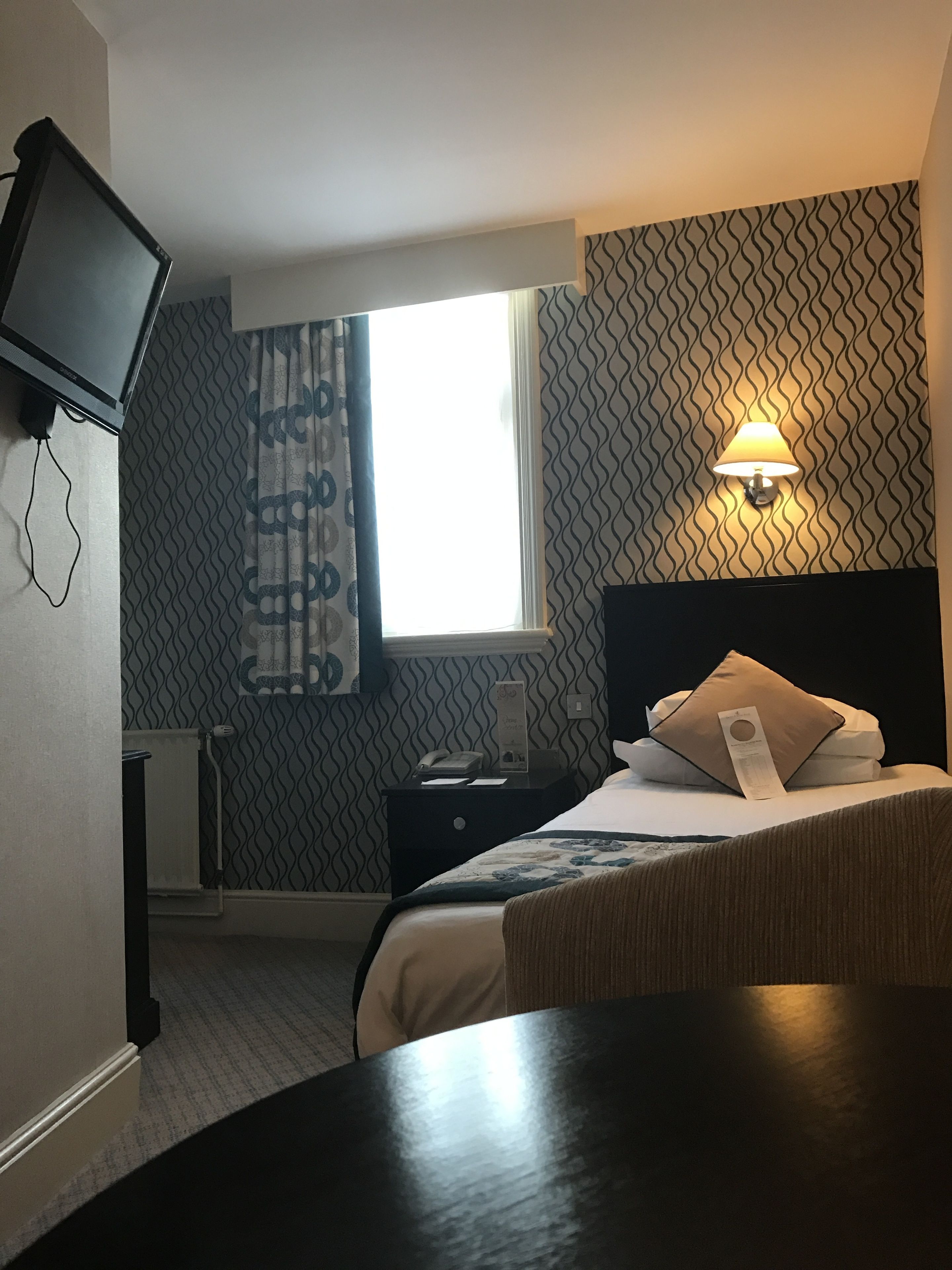 Habitación de hotel con cama, mesita de noche, lámpara, televisor montado en la pared y ventana con cortinas.