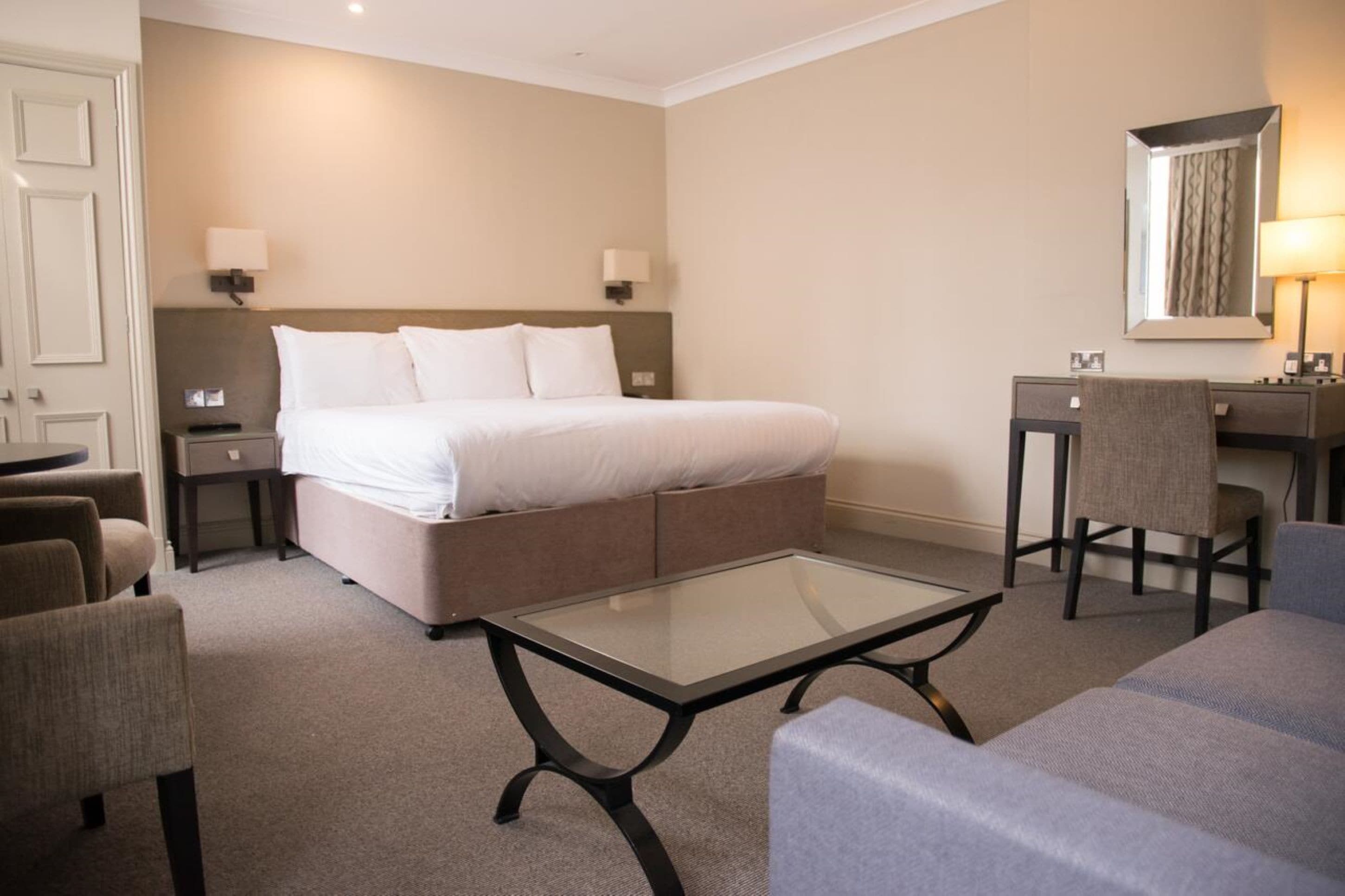 Una camera d'albergo con un letto, una scrivania, due sedie, un tavolino in vetro e uno specchio.