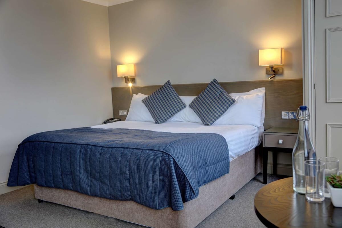 A neatly made bed with a blue blanket, two pillows, and two wall-mounted lamps.