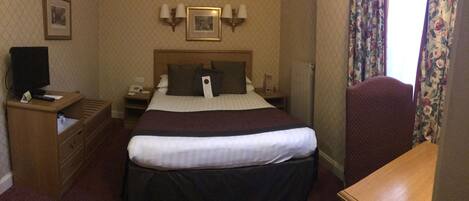Habitación de hotel con cama, escritorio, silla, televisión y mesita de noche con teléfono y lámpara.