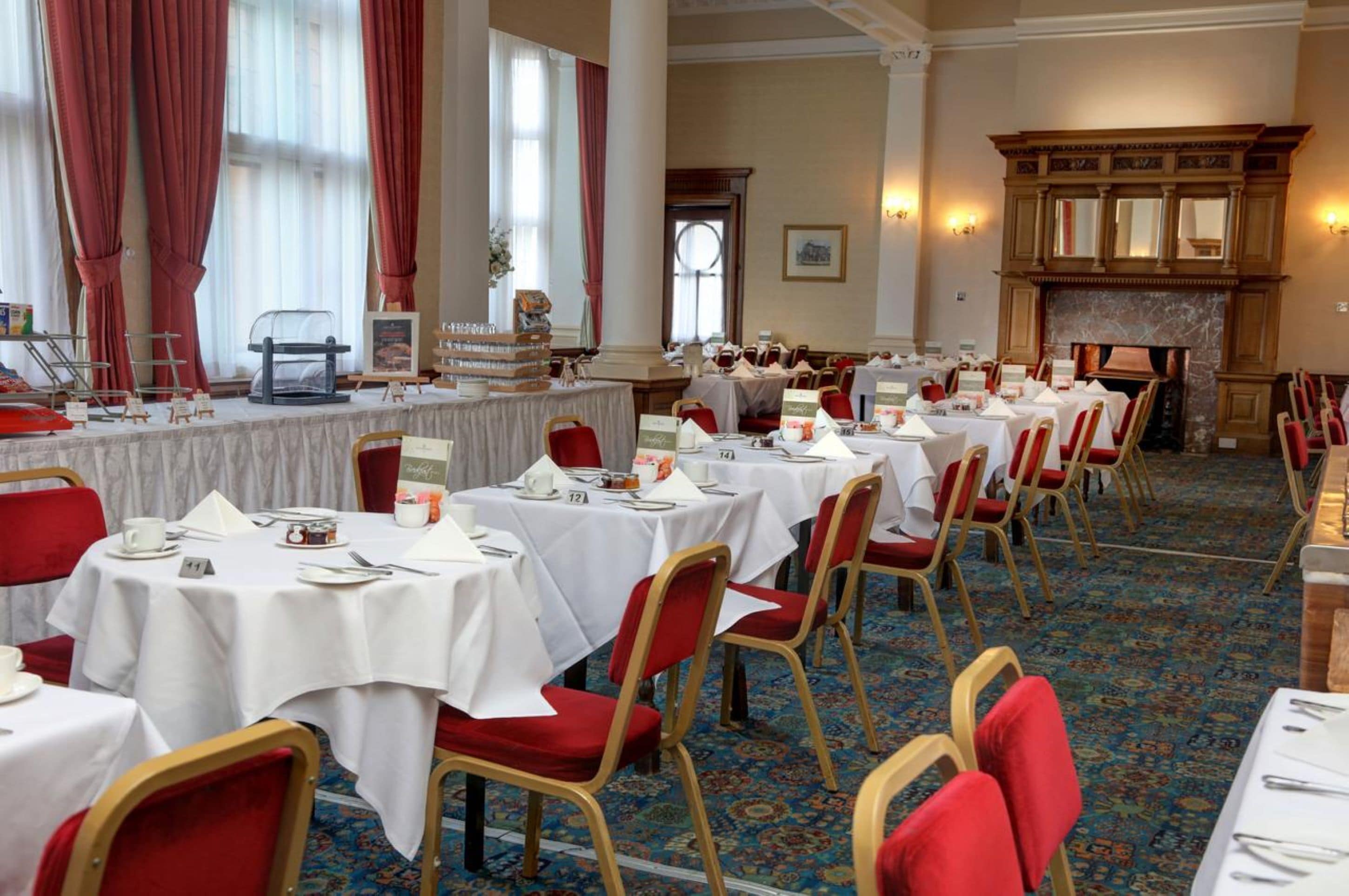 A banquet hall with multiple round tables, red chairs, and a fireplace.