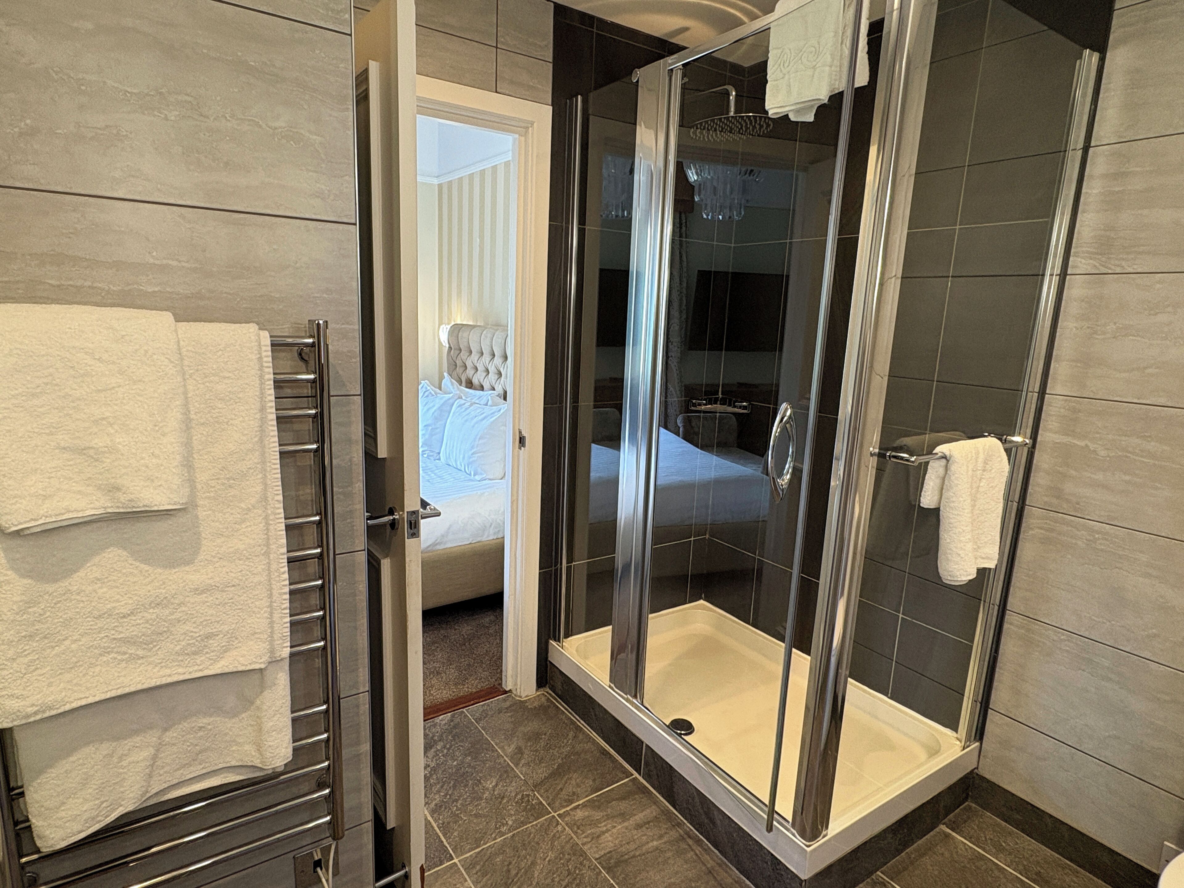 A modern bathroom with a glass-enclosed shower, a visible bed in the adjacent room, and a towel rack.