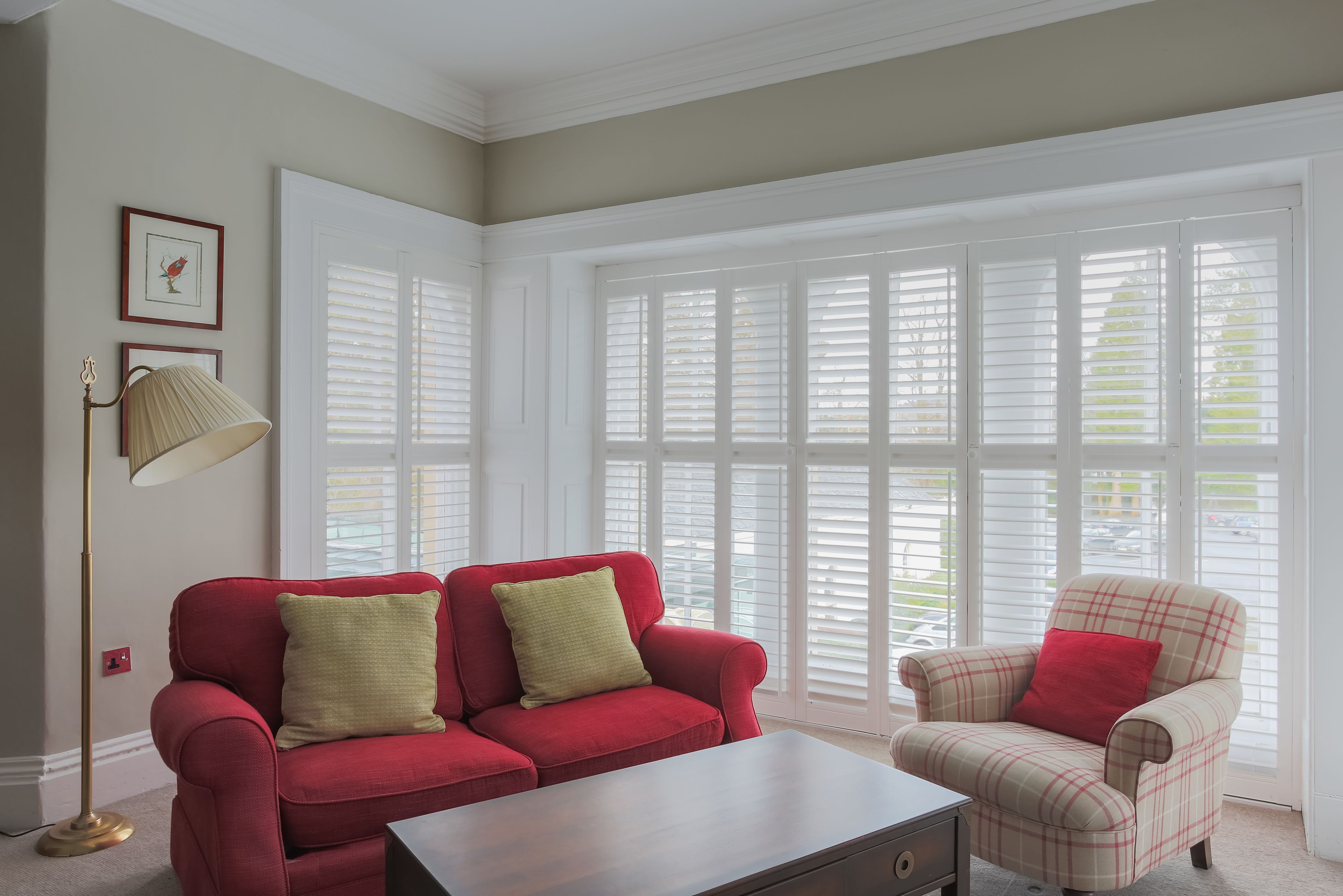 A room with a red sofa, a plaid armchair, a floor lamp, and a large window with blinds.