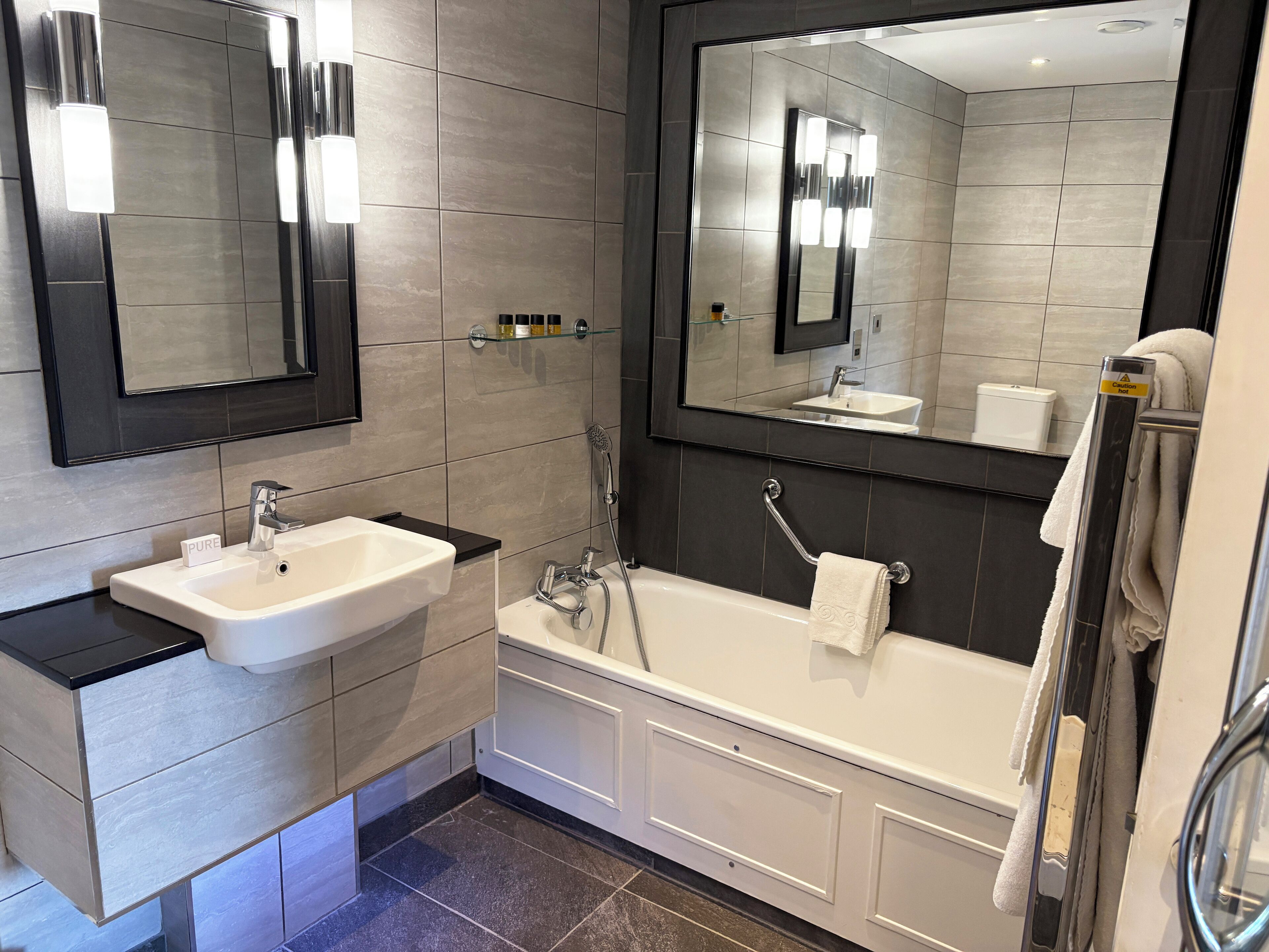 Une salle de bain moderne avec un lavabo, un meuble-lavabo et une baignoire, le tout de couleur blanche. On y trouve deux miroirs, un au-dessus du lavabo et l’autre au-dessus de la baignoire. Les murs sont recouverts de carreaux, et un porte-serviettes est placé à côté de la baignoire.