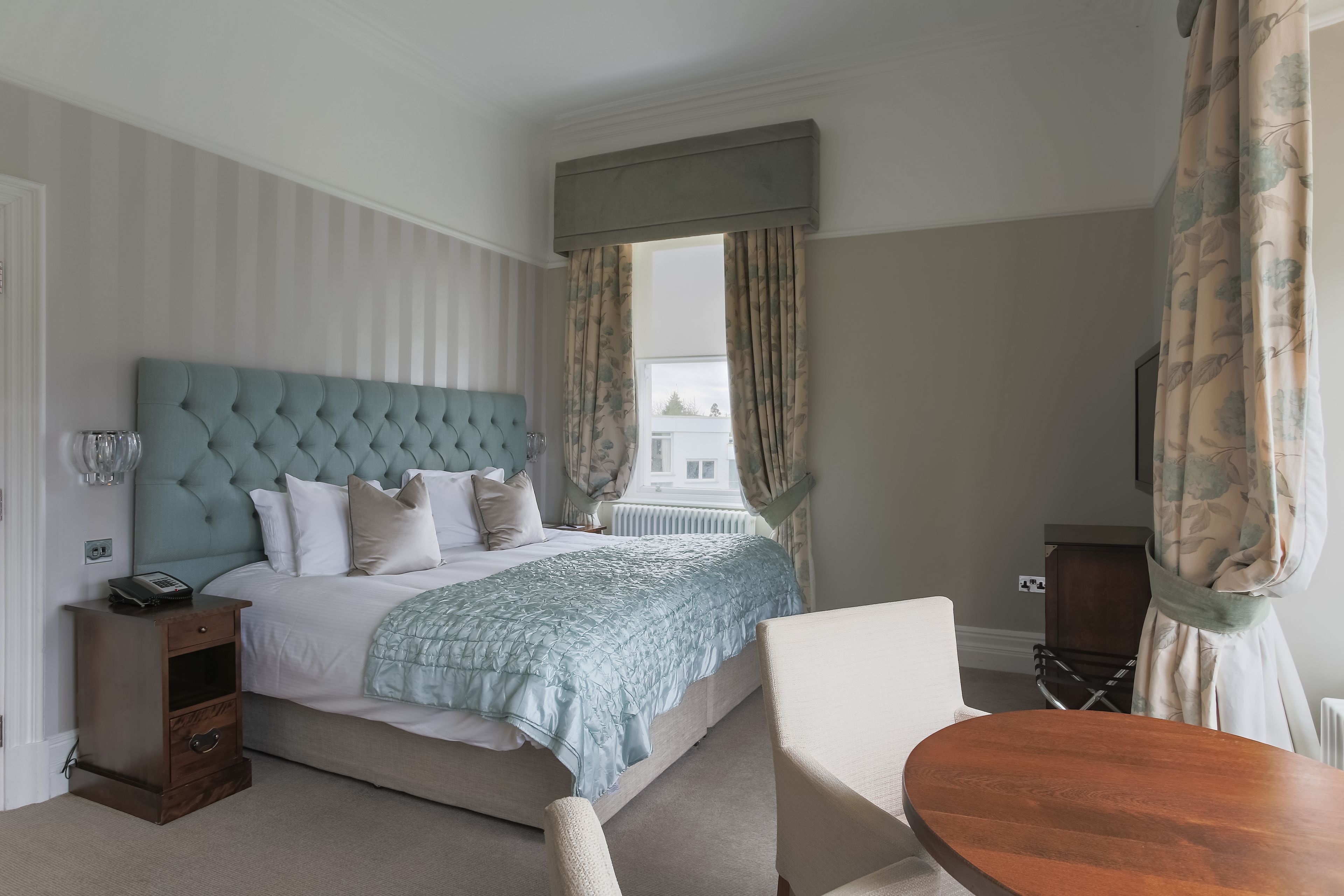 A neatly arranged bedroom with a tufted headboard, a bedside table with a lamp, and a view of the city through large windows.