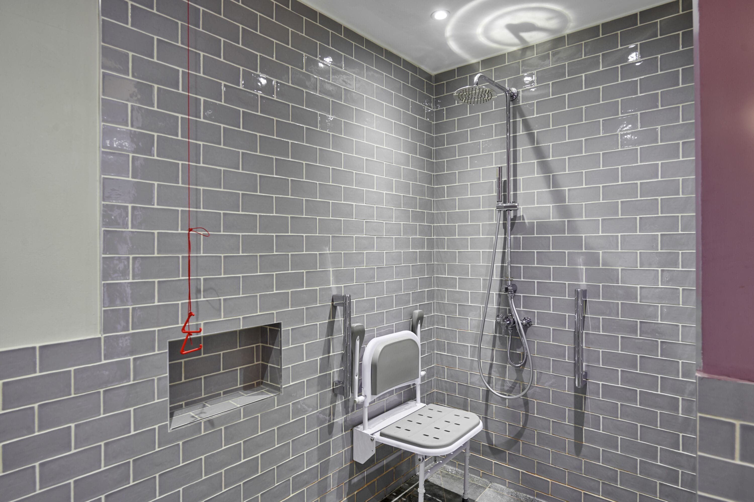 A modern bathroom with grey tiled walls, a shower with a handheld showerhead, and a white toilet with a safety bar.