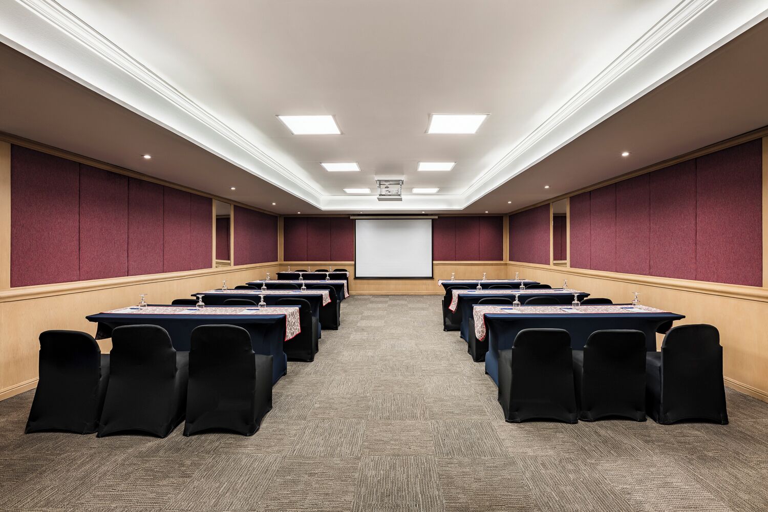 Een vergaderruimte met rijen tafels en stoelen, een scherm voor een projector en een vloerbedekking.