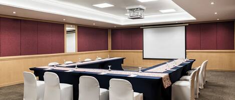 A conference room with a long table, white chairs, a projector screen, and a carpeted floor.