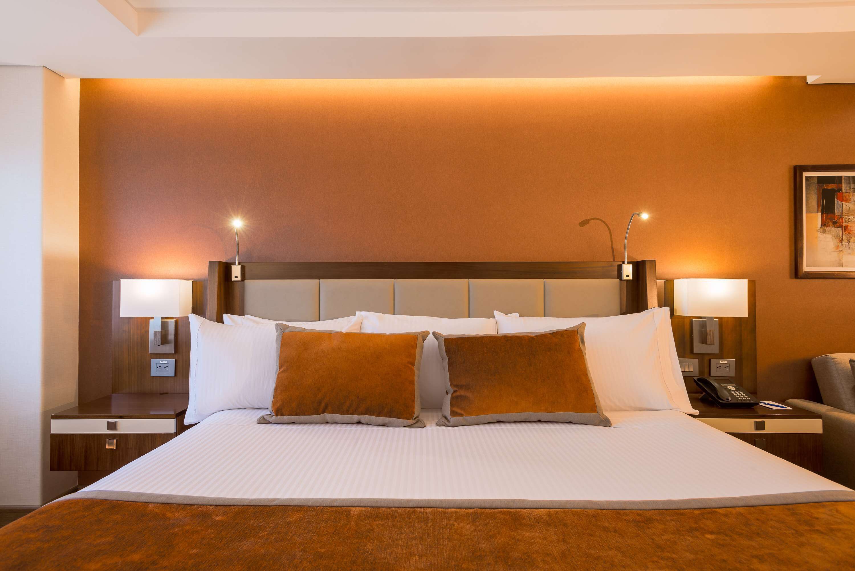 Habitación de hotel con una cama grande, dos mesitas de noche con lámparas, una silla y un cuadro en la pared.