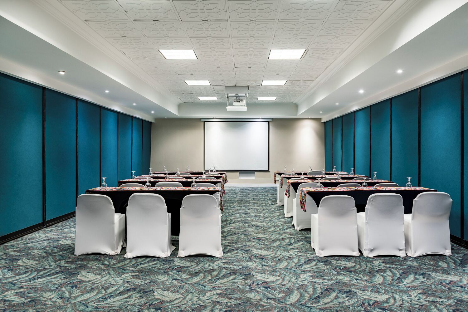 A conference room with multiple tables and chairs, a projector screen, and a patterned carpet.