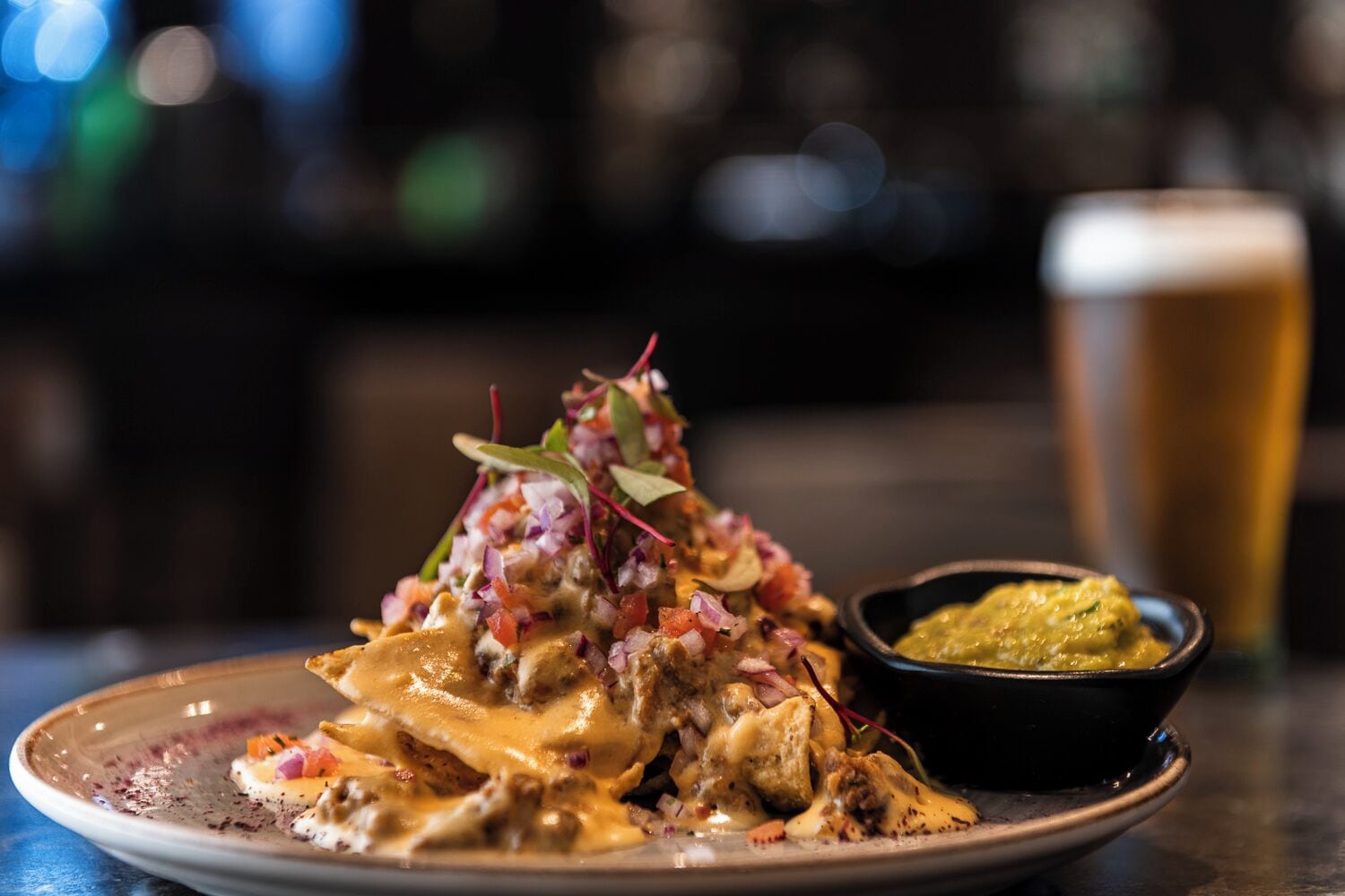 A plate of nachos topped with cheese, onions, and a green sauce, accompanied by a glass of beer and a small bowl of green sauce.