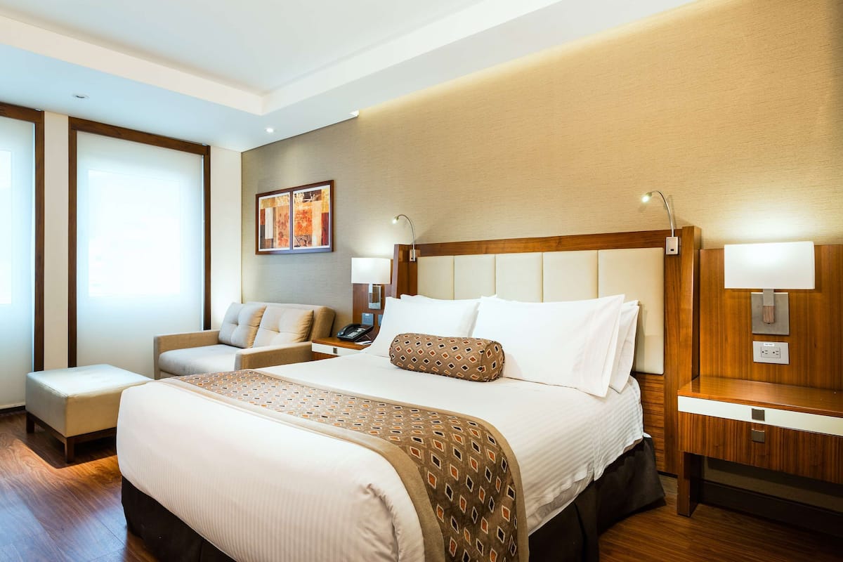 Habitación de hotel con una cama grande, una mesita de noche, una silla, un escritorio y una lámpara de pared.