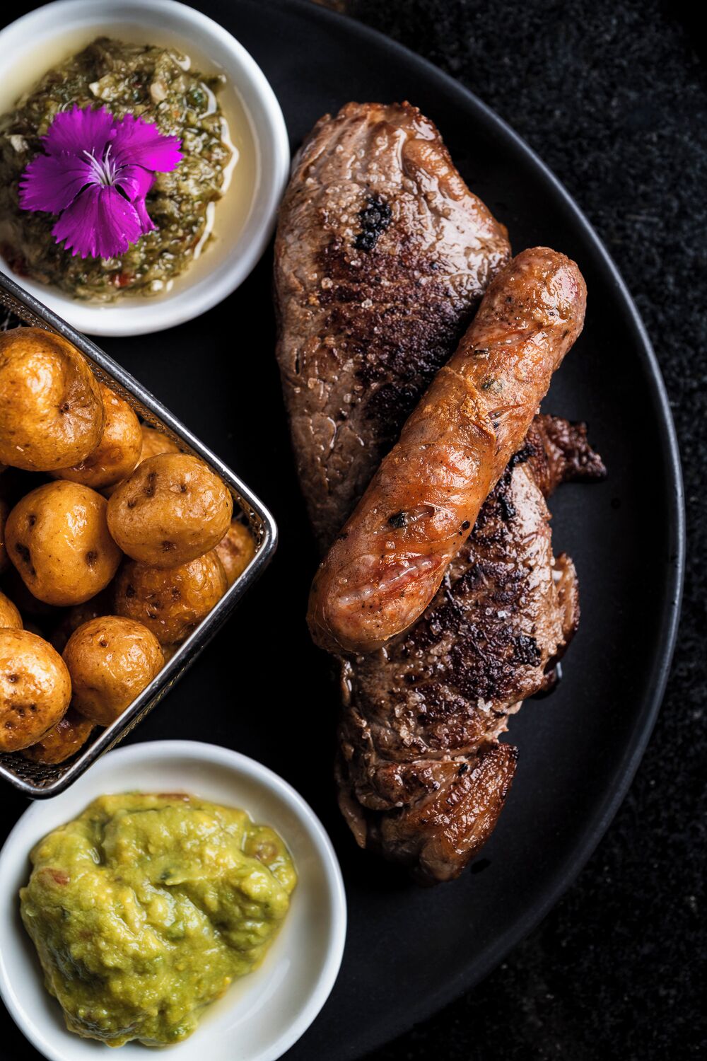 A plate with grilled meat, sausages, roasted potatoes, and a green sauce.