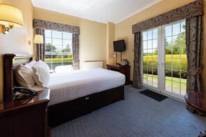 A hotel room with a large bed, a bedside table with a lamp, a TV, and a view of the garden through the window.