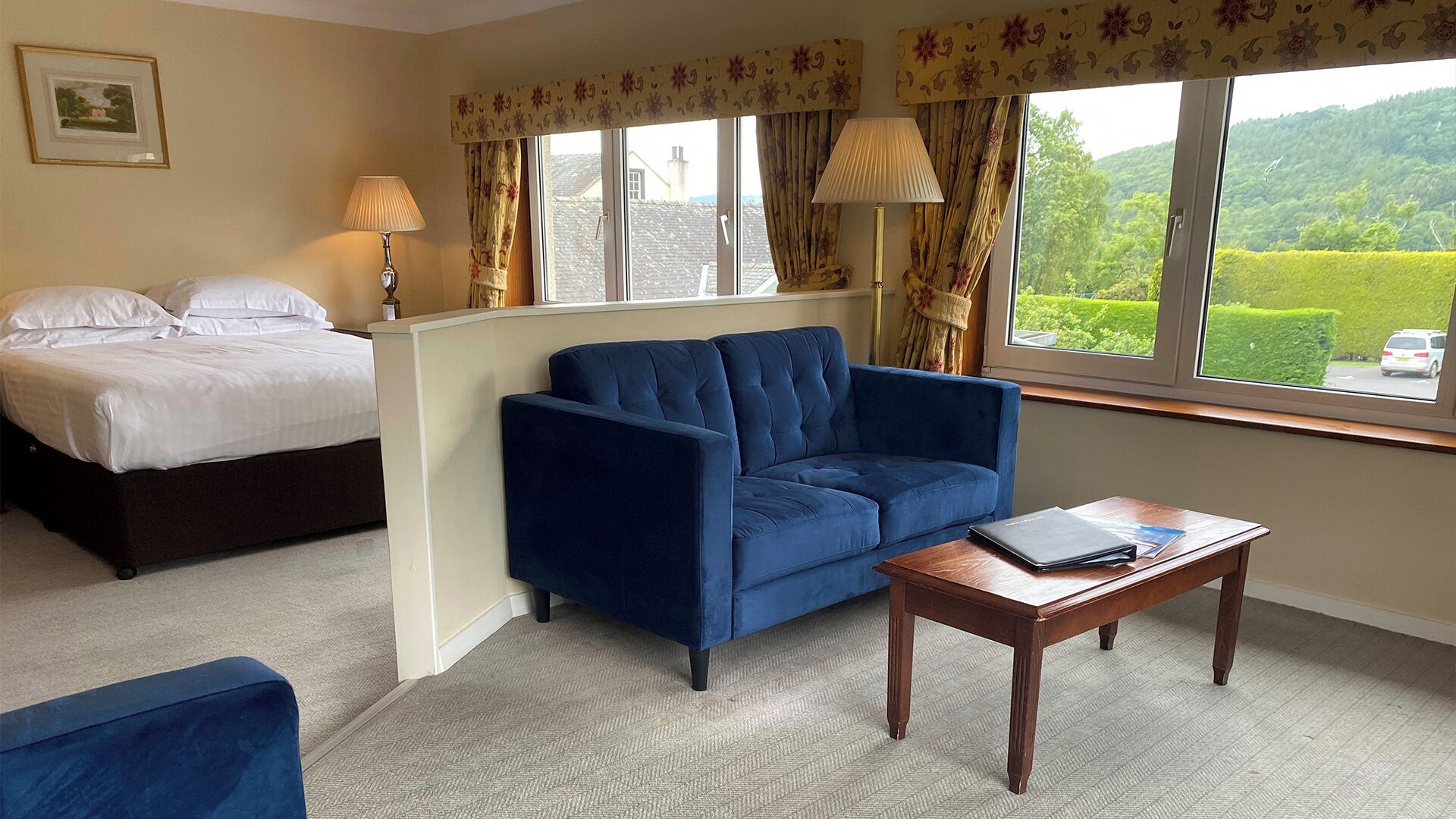 A hotel room with a bed, a blue sofa, a wooden coffee table, and a window with a view of greenery.