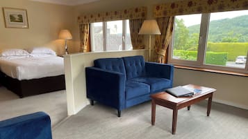 A hotel room with a bed, a blue sofa, a wooden coffee table, and a window with a view of greenery.