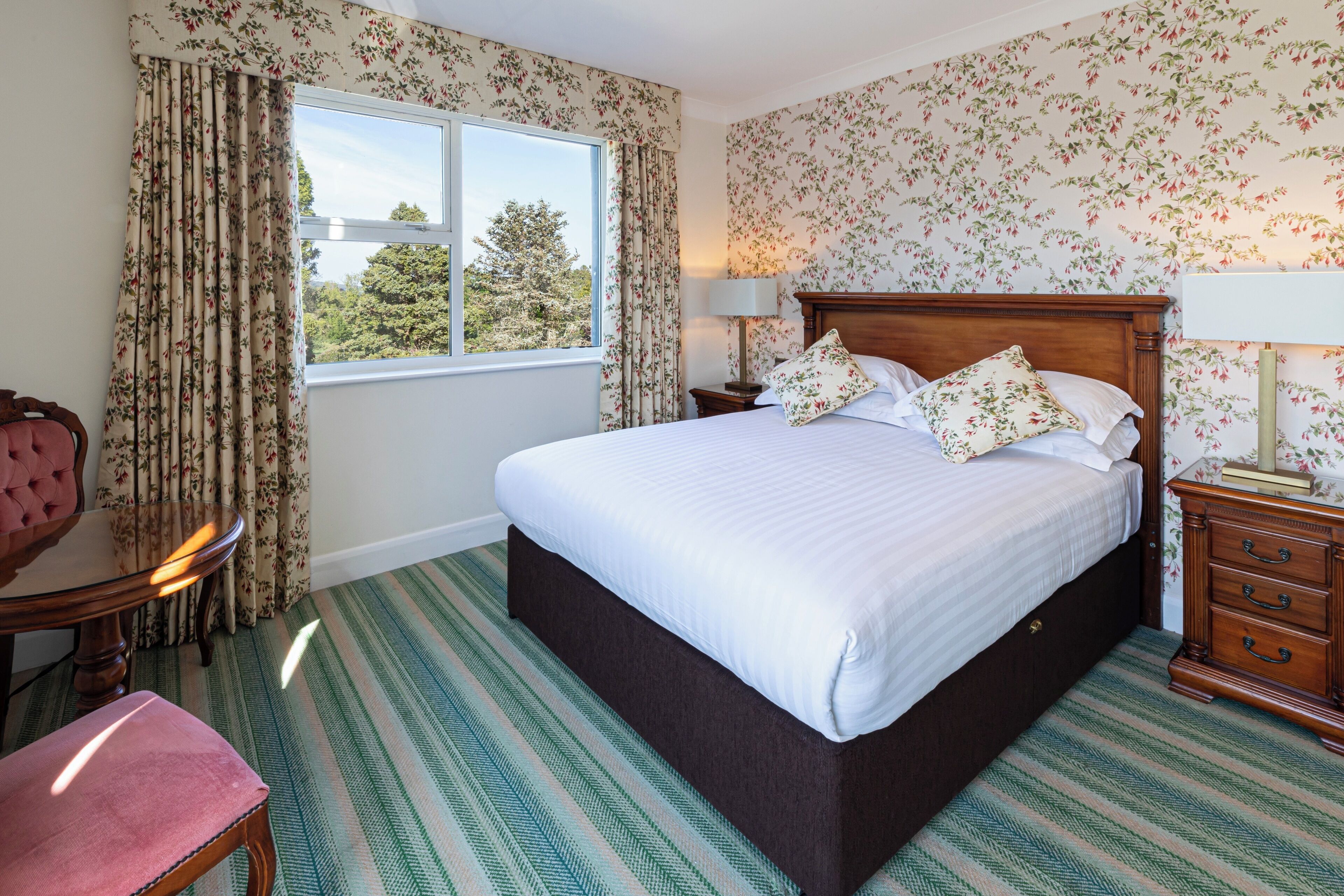 A bedroom with a large bed, floral wallpaper, a window with curtains, a bedside table with a lamp, and a chair with a cushion.