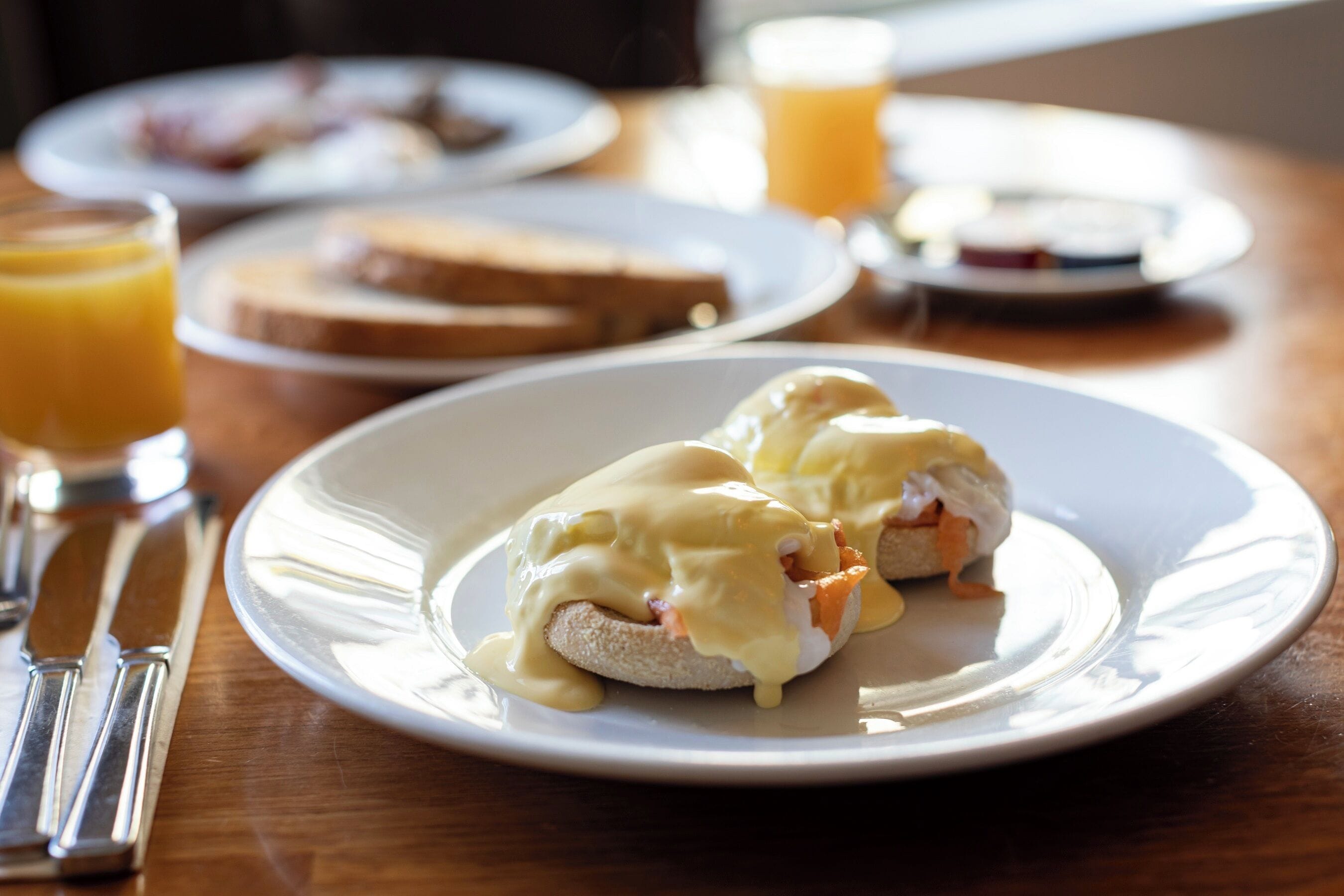 En frukostuppläggning med ägg benedict på en tallrik, rostat bröd och ett glas apelsinjuice på ett träbord.