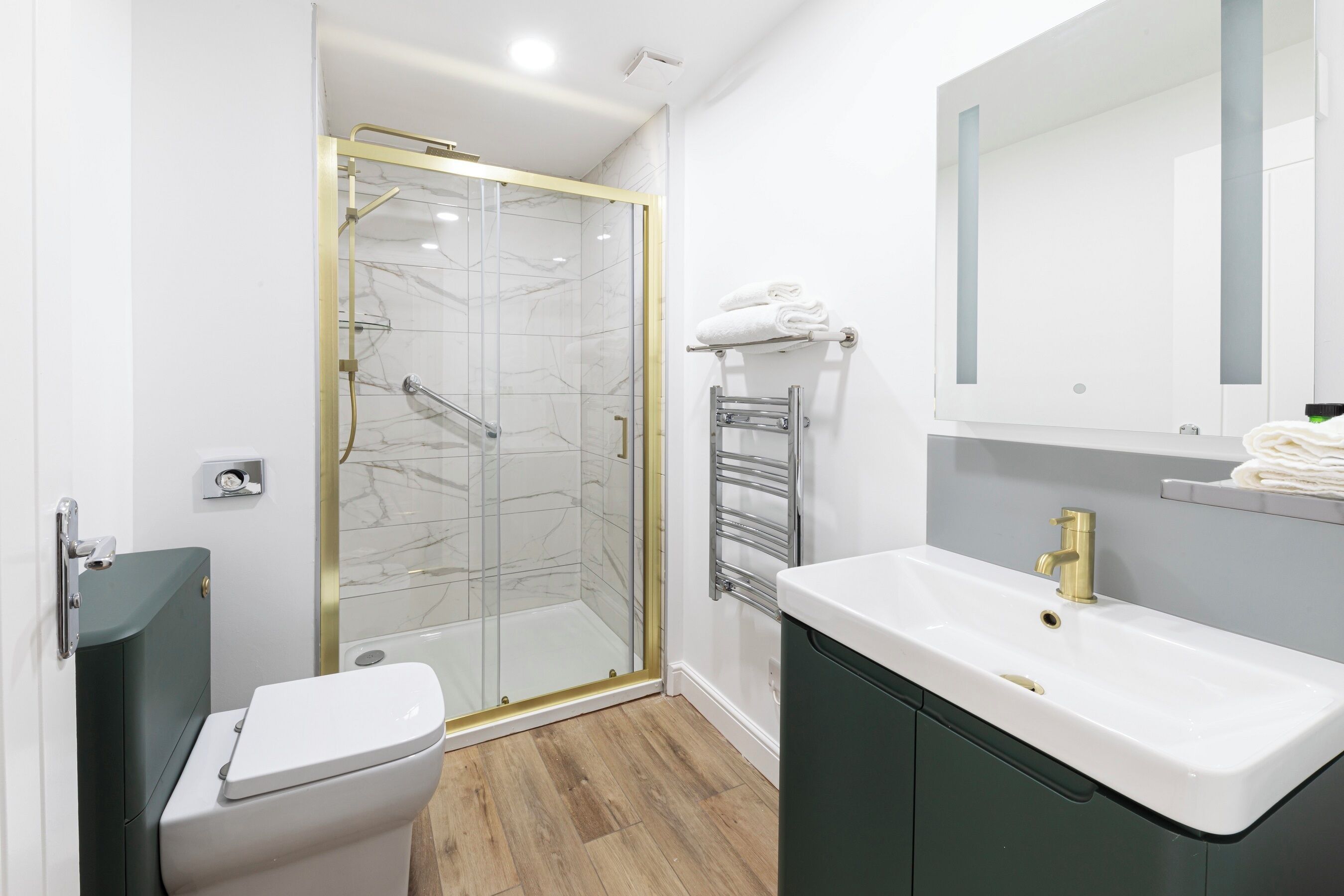 A modern bathroom with a glass-enclosed shower, a white sink with a gold faucet, and a dark green cabinet.