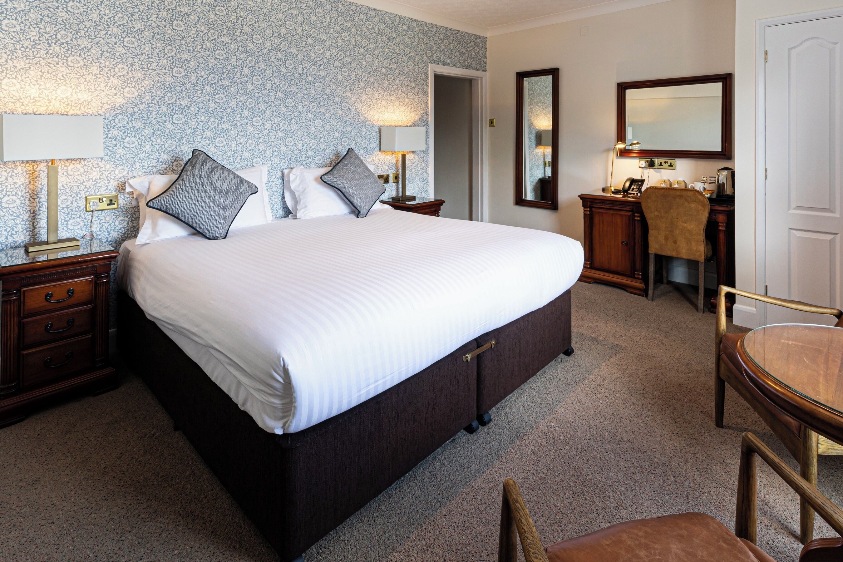 A hotel room with a large bed, bedside tables with lamps, a desk with a chair, and a mirror on the wall.