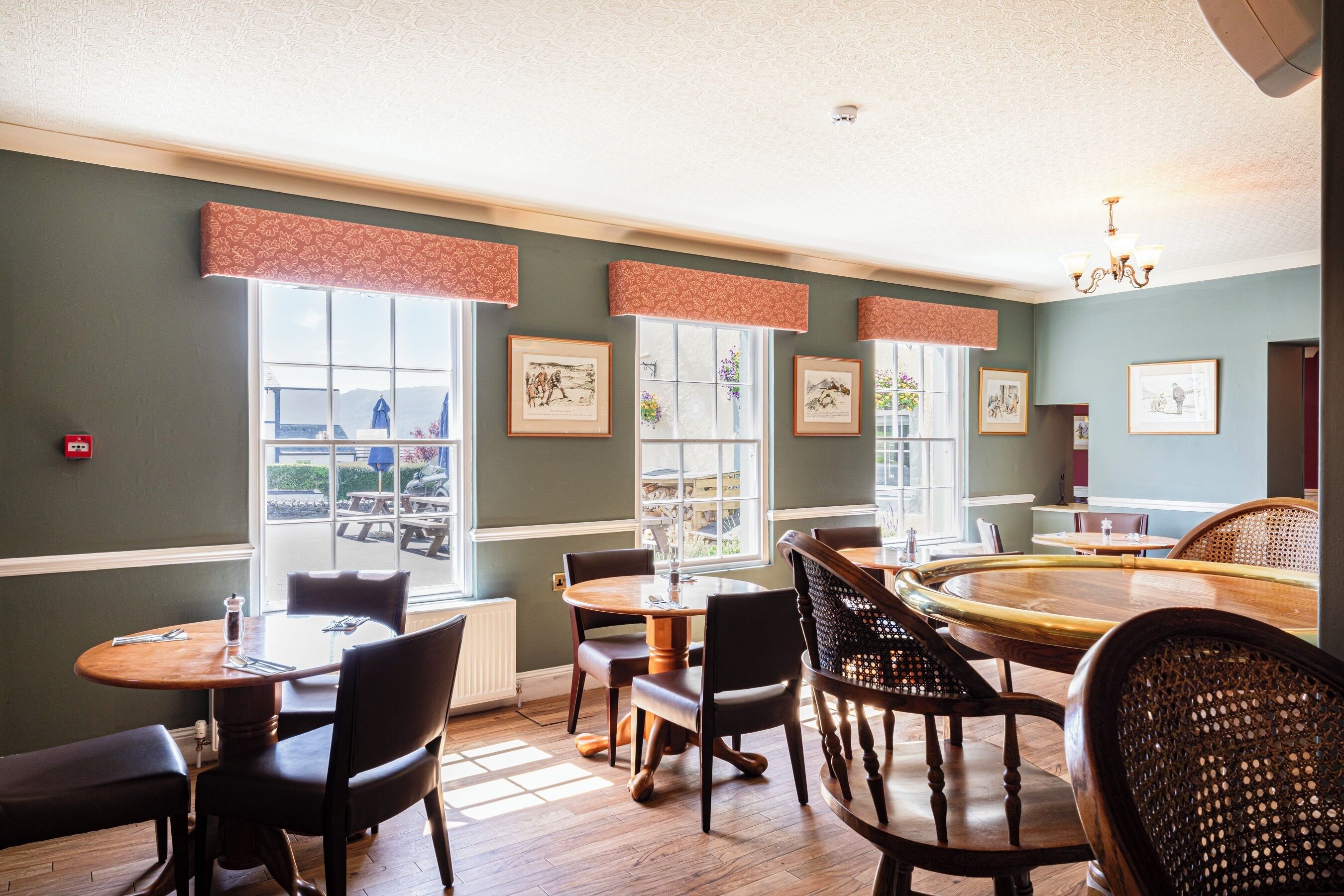 A dining area with round tables and chairs, large windows with curtains, and framed artwork on the walls.
