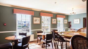 A dining area with round tables and chairs, large windows with curtains, and framed artwork on the walls.
