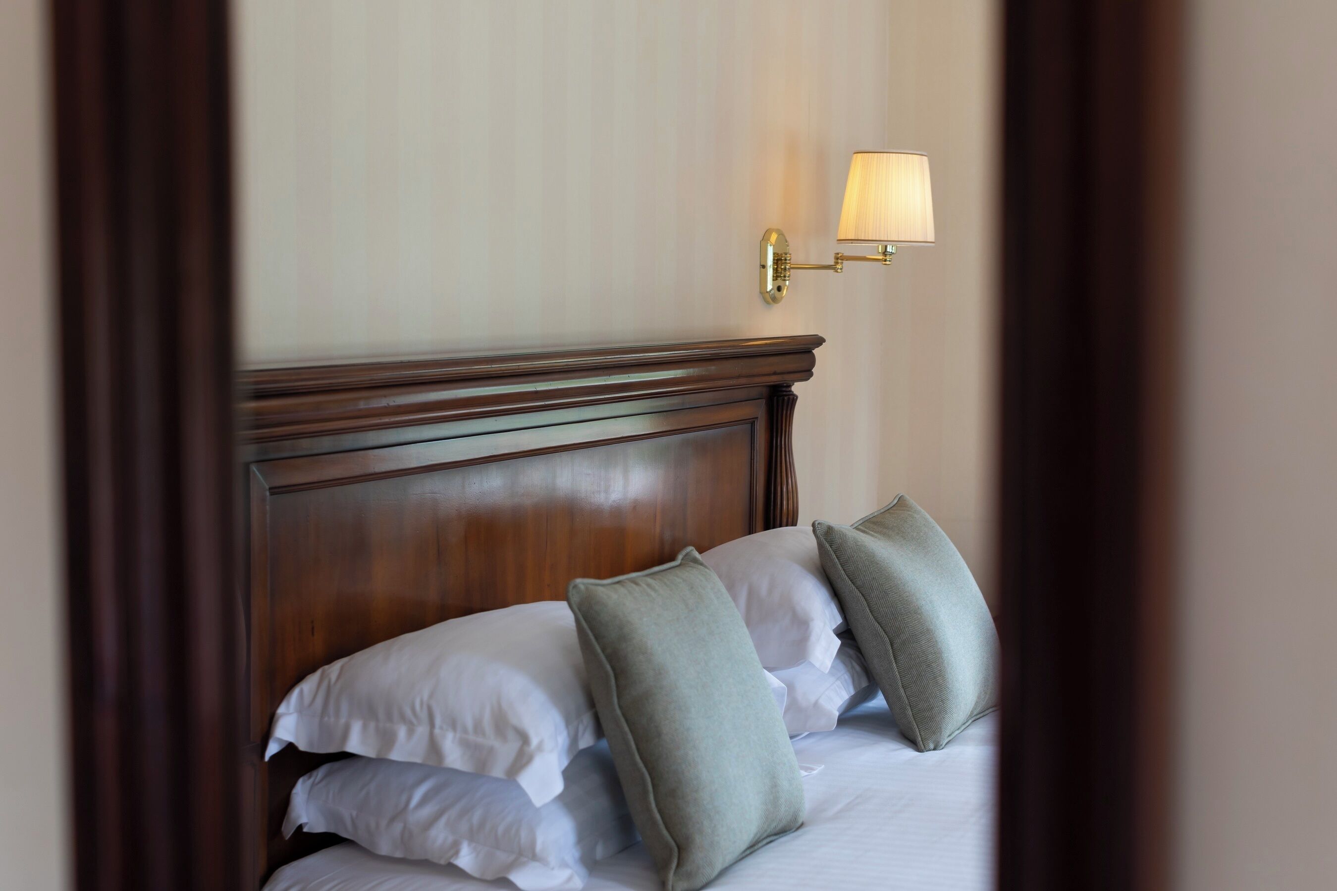 A neatly made bed with white and green pillows, a wooden headboard, and a wall-mounted lamp.