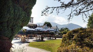 An outdoor seating area with tables and umbrellas, surrounded by greenery and a mountain in the background.