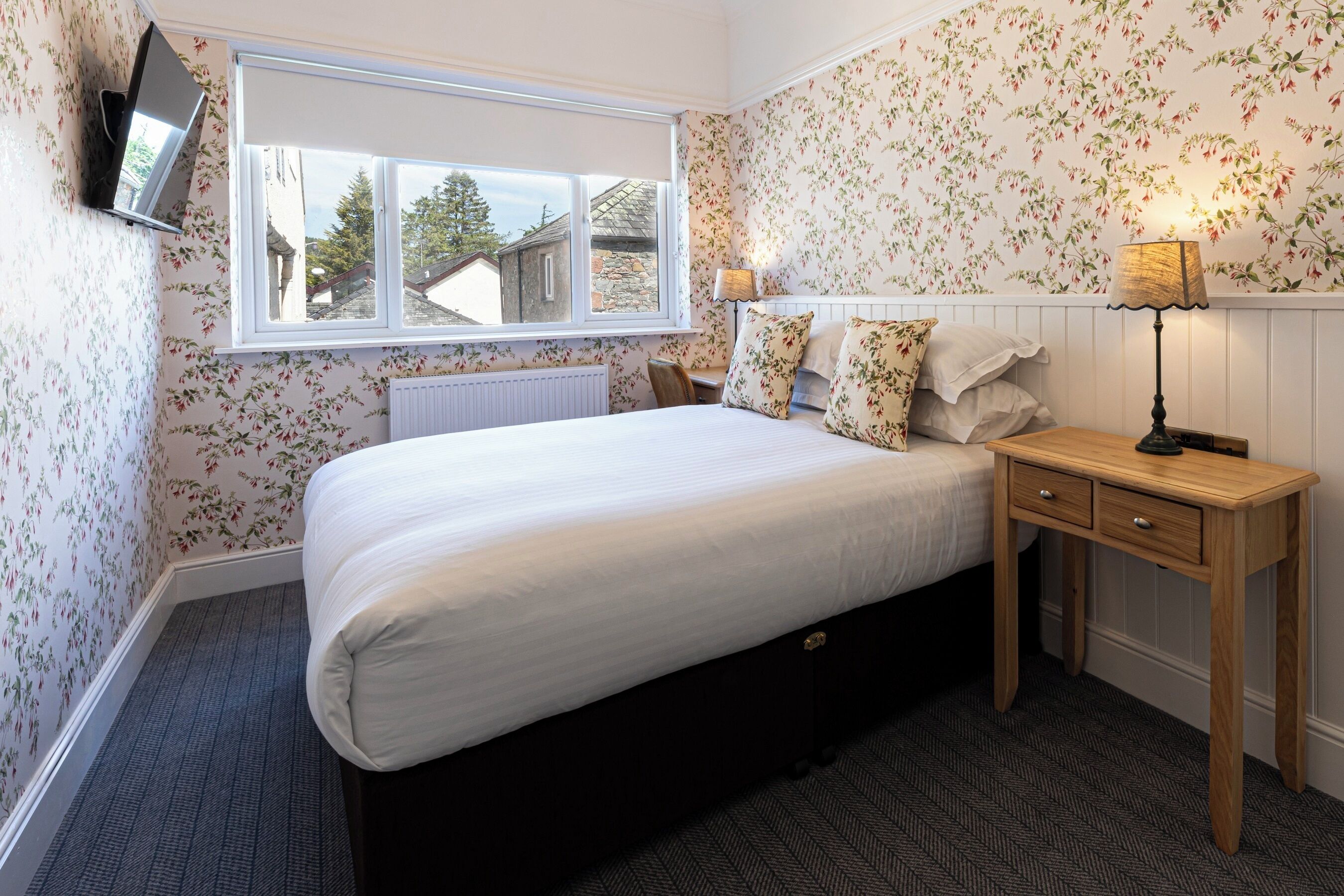 A bedroom with a large bed, a wooden nightstand, a desk, a window, and floral wallpaper.
