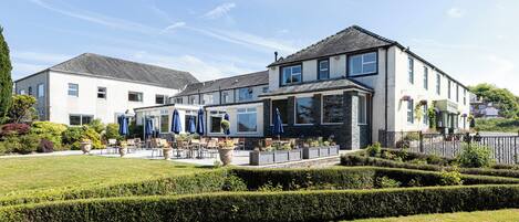 A hotel with a patio area featuring outdoor seating and umbrellas, surrounded by well-maintained gardens and hedges.