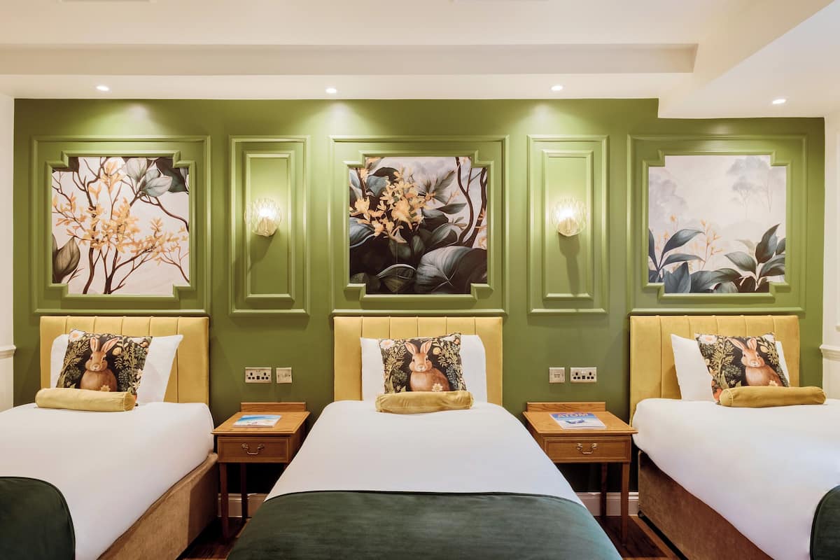 A hotel room with three beds, each with a headboard and a pillow featuring a rabbit design. The walls are painted green, and there are three wall-mounted paintings depicting floral and nature scenes.