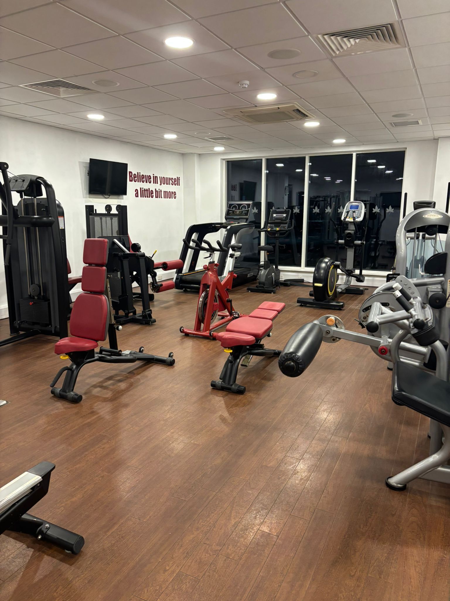 Un gimnasio con diversos equipos de ejercicio, una frase motivacional en la pared y un amplio ventanal.