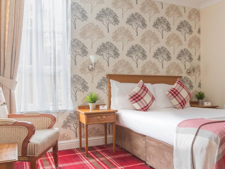 A hotel room with a bed, a chair, a nightstand, a lamp, and a window with sheer curtains.