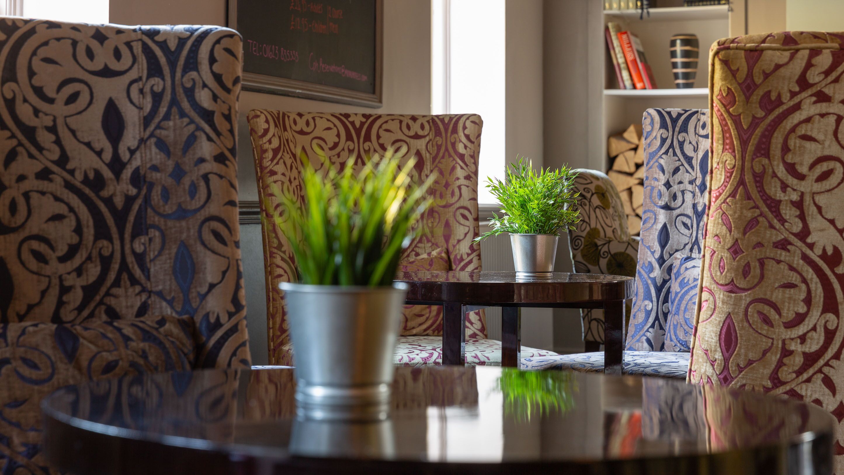 A cozy seating area with patterned upholstery, a small table with a potted plant, and a bookshelf in the background.